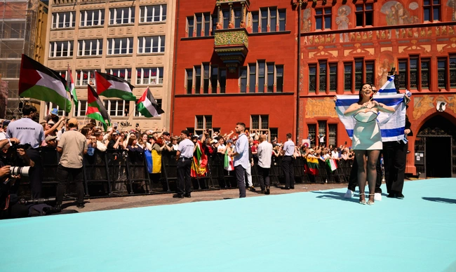 Η Yuval Raphael στο τιρκουάζ χαλί, πίσω της διαδηλωτές υπέρ της Παλαιστίνης